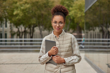 Photo of attractive curly haired student dressed in casual clothes holds modern tablet looks directly at camera poses against blurred background in city. People technology and lifestyle concept