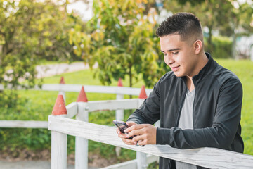 Fototapeta premium enterprising young man next to the white wooden fence with his cell phone in his hands looking at his social networks, happy for his achievements, with a beautiful sunset in the background.