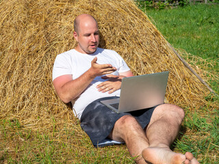 Naklejka na ściany i meble a middle-aged man, dressed in a T-shirt and shorts, is lying on a yellow haystack with a gray laptop in his hands. Remote work, training. Rural area. Indignation on the face, emotion