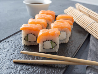 Philadelphia sushi rolls on a black textured plate stand on a gray background. There are bamboo chopsticks nearby. Side view, Japanese cuisine