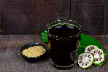 Noni juice in transparent glass and fresh noni fruit with green leaf on rustic wooden background.
