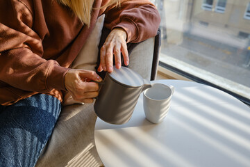 Girl pours hot tea into a cup from a teapot. Enjoy the comforts of home. White cup close-up in the hands of a woman in the rays of the sun from the window. A cup of tea in the hands of a woman