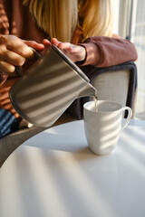 Girl pours hot tea into a cup from a teapot. Enjoy the comforts of home. White cup close-up in the hands of a woman in the rays of the sun from the window. A cup of tea in the hands of a woman