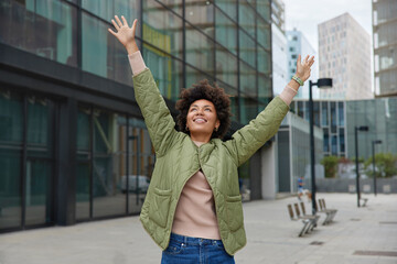 Horizontal shot of happy carefree woman with curly hair keeps arms raised up enjoys life poses during daytime in city wears jacket and jeans looks overhead has cheerful expression. People and freedom