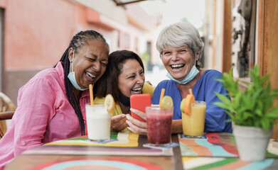 Elderly multiracial women having fun at bar outdoor using mobile phone while wearing safety mask under chin