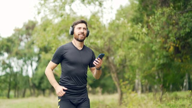 young cheerful active guy sporty running with headphones mobile phone enjoying listening to music, jogging outdoors city park or forest background. Man runner makes fitness workout earphones