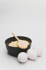 dark stoneware bowl with flour wooden spoon and white eggs on a white tabletop