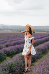 A young beautiful girl in a delicate dress and hat walks through a beautiful field of lavender and enjoys the aroma of flowers. Vacation and beautiful nature.