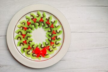 Fruit Christmas platter wreath made it from kiwi fruit,pomegranate and red bell pepper on plate with white wood background.Healthy vegan art food idea for Christmas party.Top view.Copy space