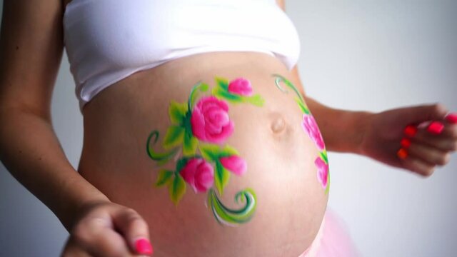 Close-up of the belly of a dancing pregnant woman on a white background, face painting of pink flowers with green petals is drawn on the belly. High quality FullHD footage