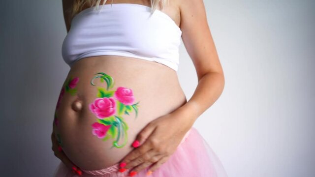 Close-up of the belly of a dancing pregnant woman on a white background, face painting of pink flowers with green petals is drawn on the belly. High quality FullHD footage
