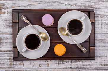 Two cups of coffee, spoon and macaron cakes on tray on white wooden table