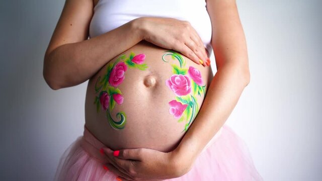 A close-up of the abdomen of a pregnant woman on a light background, the girl strokes the pregnant stomach. On a pregnant belly painted aqua makeup pink flowers with green leaves. High quality FullHD