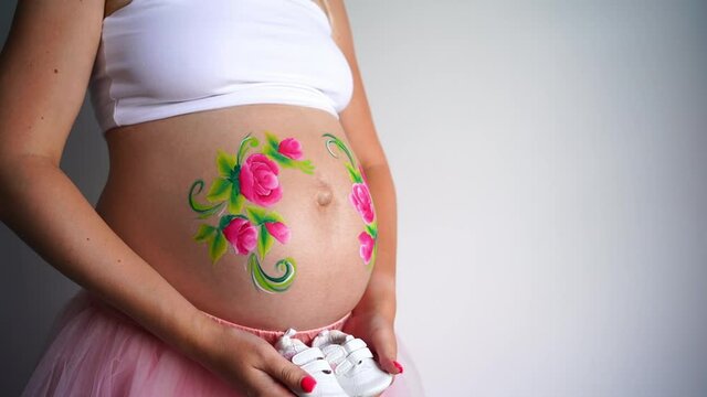 A close-up of the abdomen of a pregnant woman on a white background, on the stomach painted aqua makeup of pink flowers with green petals. A girl near a pregnant belly in her hands holds white booties