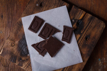 Broken bar of dark bitter chocolate with chilly pepper and red pea pepper inside on white paper, wooden board background. Unusual sweets taste