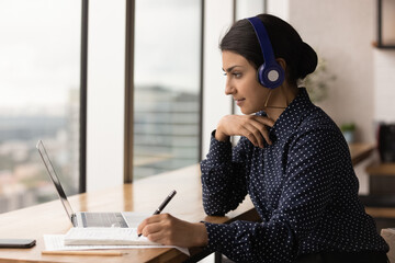 Concentrated young indian woman in headphones looking at computer screen, improving knowledge...