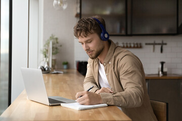Happy young handsome man in headphones listening educational lecture, handwriting notes, enjoying improving knowledge holding video call conversation with teacher or studying on online courses.