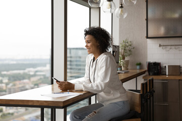 Dreamy beautiful happy young african american woman freelancer holding smartphone in hands, sitting at table in modern kitchen near panoramic window with cityscape view, planning distant workday.