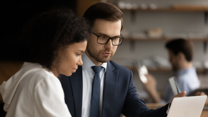 Serious smart young male caucasian team leader coaching african american female employee, explaining computer software applications, working on online project or analyzing sales together in office.