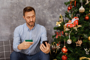 man is sitting near the Christmas tree with a mobile phone, smartphone and credit card in hands, he...