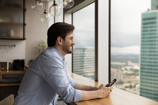 Side View Dreamy Happy Young Caucasian Man Holding Cellphone In Hands, Looking In Distance Out Of Window, Sitting At Table In Modern Kitchen, Visualizing Future, Daydreaming Or Planning Workday.