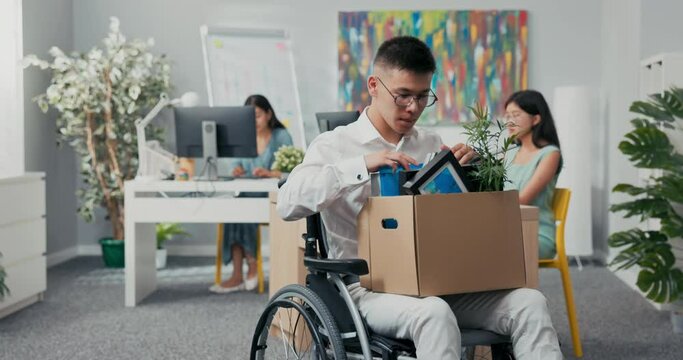 Young corporate office worker in glasses injured, accident, disabled, in wheelchair, holding a box of paraphernalia from his desk in lap, retiring, laid off, unemployed, changing positions