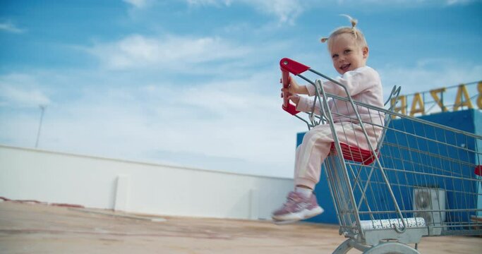 Cute child baby girl is sitting outdoors in supermarket shopping cart and smiling. Happy child waiting for parents on parking inside market troller