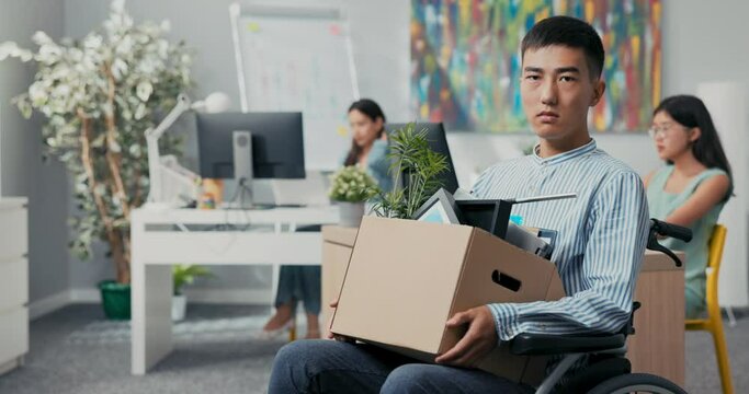 Disabled sad men in shirt with Asian Korean beauty sits in wheelchair at desk in the company on knees holding cardboard box with packed things, gets fired from job unable to fulfill his duties