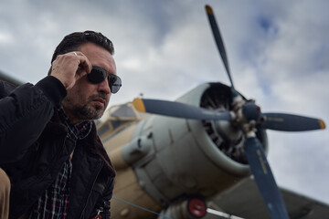 Oldschool pilot in the bomber jacket posing near the aircraft, aviator male with scarf and...