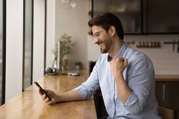 Side view happy emotional young caucasian man celebrating online lottery gambling auction giveaway win, getting victory notification on cellphone, feeling joyful sitting at table in modern kitchen.