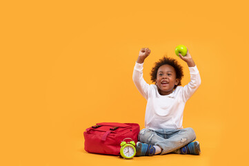 Black child boy 3 years, Student kid holding green apple in his hand and Yay! happy while sitting with school bag and alarm clock. Isolated portrait on yellow background with copy space.