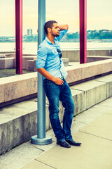 Obraz premium Man Thinking Outside. Wearing a light blue shirt, blue jeans, leather shoes, sunglasses hanging on shirt, a young guy with a little beard, mustache is leaning on a pole, sad, frowned, thinking.