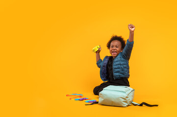 Black child boy 3 years, Student kid holding an alarm clock in his hand and Yay! happy while sitting with school bag. Isolated portrait on yellow background with copy space. International students