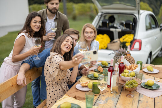 Pleased european friends taking selfie on mobile phone during friendly picnic. Young men and women drinking wine from glasses. People enjoying time together. Concept of friendship. Idea of leisure