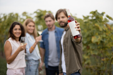 Joyful european friends tasting local wine near vineyards in countryside. Man showing a bottle with blank label to copy paste. Young men and women enjoying time together