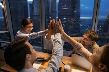 Happy young mixed race employees colleagues teammates joining hands in air, celebrating shared...