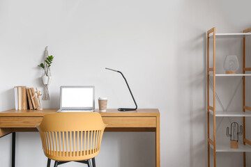 Workplace with modern laptop, lamp and paper cup of coffee on wooden table near white wall