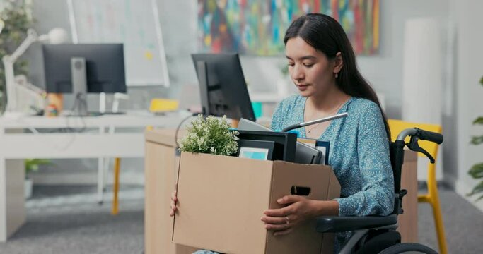 A disabled attractive woman dressed as an Asian Korean beauty sits in a wheelchair at a corporate desk on her lap holding a cardboard box of packed belongings, fired from job, thrown out of position