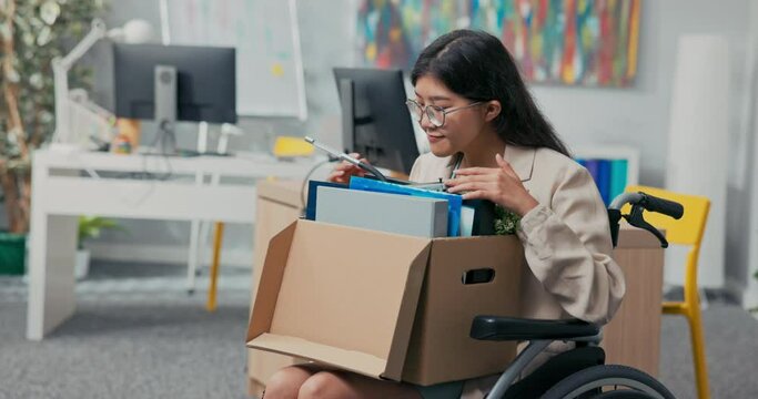 Disabled woman with glasses and Asian Korean beauty sits in wheelchair at corporate desk on lap holding a cardboard box of packed accessories, changing positions, getting promoted