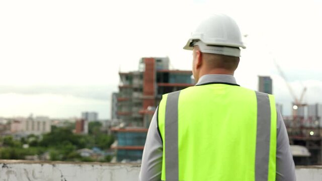 Consult engineer handsome man or architect looking and walking with white safety helmet in city construction site . Checking with checklist on rooftop building construction at capital.