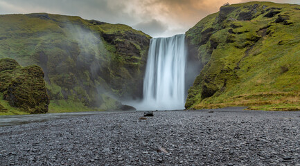 Closeup of Skogafoss waterfall in Iceland at sunrise.  Black and gray gravel in foreground. Green cliffs, no people.
