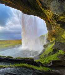 Rear cave view of Seljalandsfoss waterfall in Iceland. Cave rim has highlights or orange and green.  Patches of light on waterfall from late afternoon sunlight.