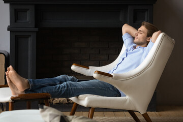 Serene young man put hands behind head, feet on footstool, relaxing in leather comfortable armchair in luxury home with fireplace. No stress, fatigue relieve, furniture store ad, peace of mind concept - obrazy, fototapety, plakaty