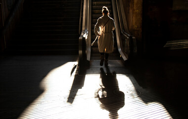 One young woman in coat walking alone up the escalator public stairs in backlight sunlight with shadows