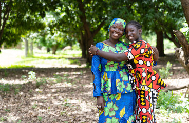 Cheerful black African teenager hugging her mother both standing in a forest among mango trees;...
