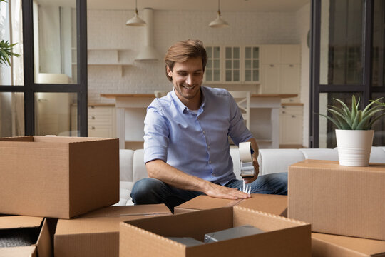 Man prepare personal stuff for relocating to own house, sit on sofa using adhesive scotch, preparing belongings for transportation, happy client of trustworthy transporting company, moving day concept