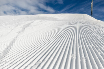 Close-up straight line rows of freshly prepared groomed ski slope piste with bright shining sun and clear blue sky background. Snowcapped mountain downhill landscape at europe winter skiing resort