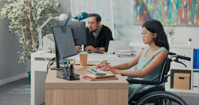 Workday in office, corporation, company, co-workers are working on computers, drafting documents, woman in glasses with Asian beauty is sitting at desk in wheelchair, she is disabled, after accident