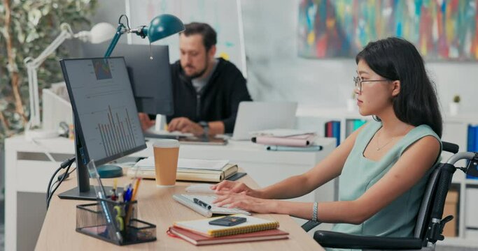 Workday in office, corporation, company, co-workers are working on computers, drafting documents, woman in glasses with Asian beauty is sitting at desk in wheelchair, she is disabled, after accident