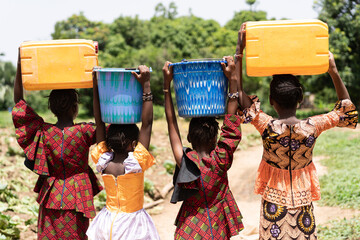 Close up rear view of black African girls carrying heavy water containers on their heads; child...